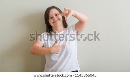 Down syndrome woman standing over wall smiling making frame with hands and fingers with happy face. Creativity and photography concept.