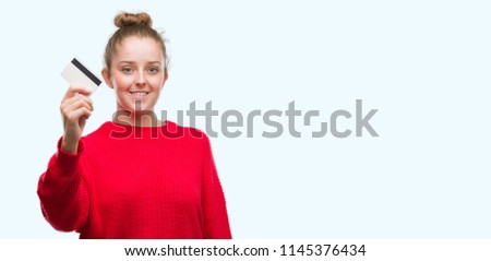 Young blonde woman holding credit card with a happy face standing and smiling with a confident smile showing teeth