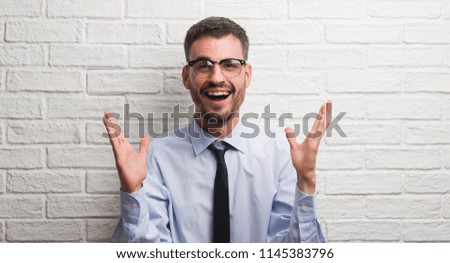 Young adult business man standing over white brick wall very happy and excited, winner expression celebrating victory screaming with big smile and raised hands
