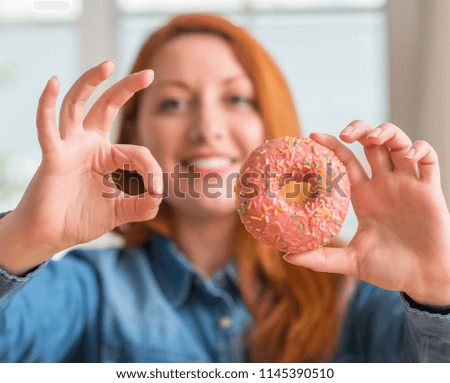 Redhead woman holding donut at home doing ok sign with fingers, excellent symbol