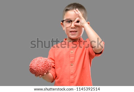 Dark haired little child with glasses holding a brain with happy face smiling doing ok sign with hand on eye looking through fingers