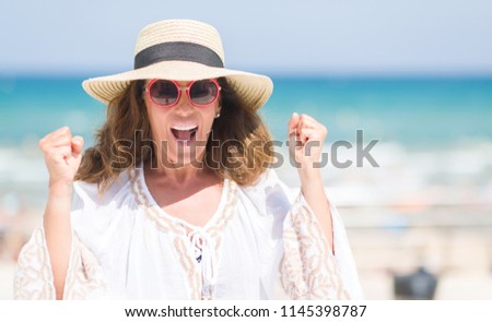 Middle age brunette woman wearing summer hat on vacations by the beach screaming proud and celebrating victory and success very excited, cheering emotion