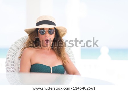 Middle age brunette woman sitting at the table by the beach scared in shock with a surprise face, afraid and excited with fear expression