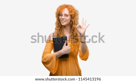 Young redhead student woman holding a book doing ok sign with fingers, excellent symbol
