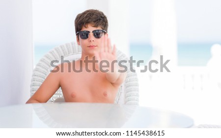 Young child on holidays sitting on a chair by the beach with open hand doing stop sign with serious and confident expression, defense gesture