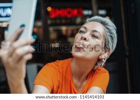 Attractive blonde girl in orange T-shirt making selfie at cafe and showing her tongue
