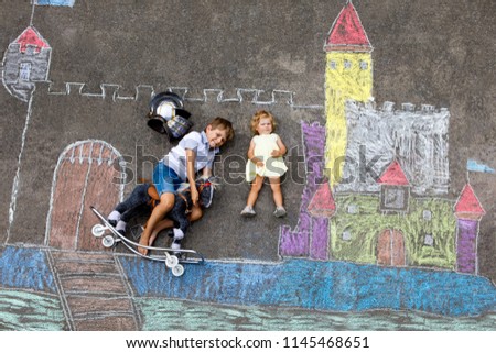 Little active kid boy and cute toddler baby girl drawing knight castle and fortress with colorful chalks on asphalt. Happy children with helmet and rocking horse toy having fun with playing knight.