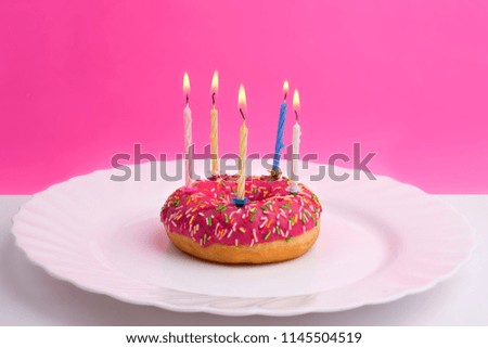 Donut in the form of a birthday cake with burning candles on a white plate on a bright pink background