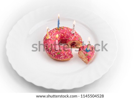 Donut in the form of a birthday cake with burning candles and a cut piece of cake with a candle on a white plate on a white background top view