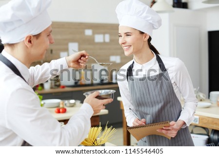 Smiling beautiful young woman in chefs hat and apron tasting food of intern cook, young man giving spoon with sauce to examiner