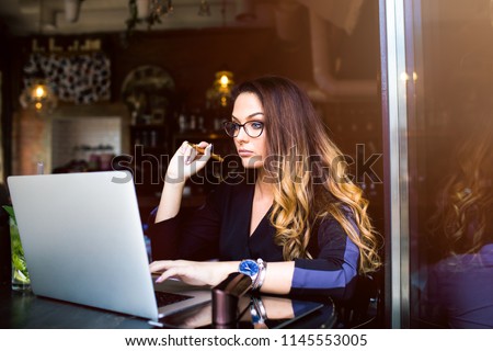 Gorgeous woman university student learning online via laptop computer, sitting in cozy coffee shop. Female in stylish glasses skilled freelance social media content writer working on pc notebook
