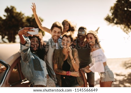 Group of young men and women enjoying on road trip standing by the car and taking selfie. Multiracial group of friends having fun outdoors taking selfie with smart phone.