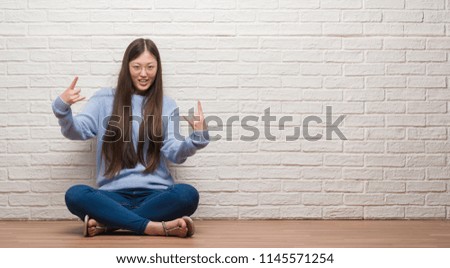 Young Chinese woman sitting on the floor over brick wall shouting with crazy expression doing rock symbol with hands up. Music star. Heavy concept.