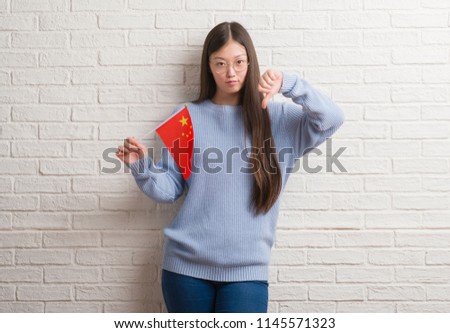 Young Chinese woman over brick wall holding flag of China with angry face, negative sign showing dislike with thumbs down, rejection concept
