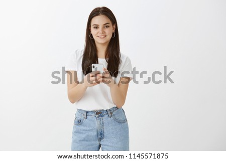 What is your profile nickname again. Stylish modern woman in trendy clothes, holding smartphone and gazing with happy expression at camera, waiting to take picture on phone, standing over grey wall