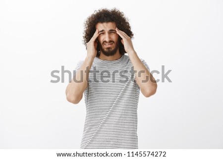 Prtrait of questioned unfocused cute male in striped t-shirt, squinting and holding hands on temples, trying to concentrate, having headache or migraine, standing confused over gray background