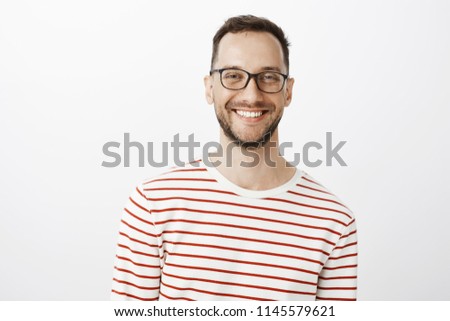 Waist-up shot of relaxed good-looking positive guy in glasses, smiling broadly and gazing at camera while talking with friend, standing against gray background, discussing interesting topic
