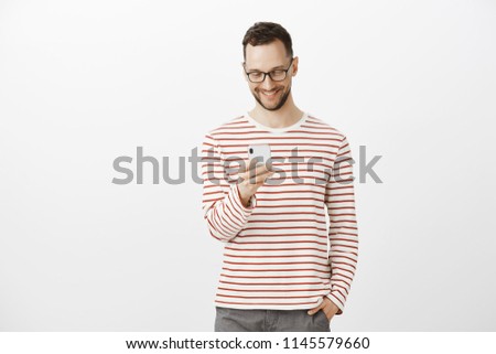 Pleased carefree handsome man with bristle in glasses, holding hand in pocket while brosing in net via smartphone, smiling joyfully at screen, enjoying great quality of cellphone over gray wall