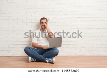 Young caucasian man sitting over white brick wall using computer laptop with happy face smiling doing ok sign with hand on eye looking through fingers