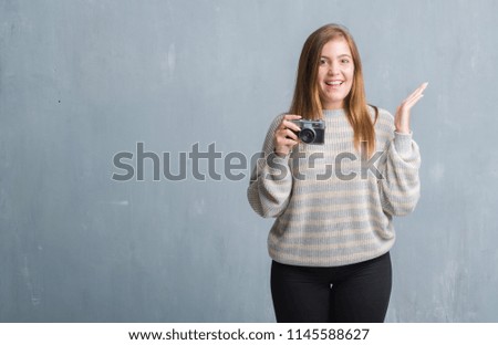 Young adult woman over grey grunge wall taking pictures using vintage camera very happy and excited, winner expression celebrating victory screaming with big smile and raised hands