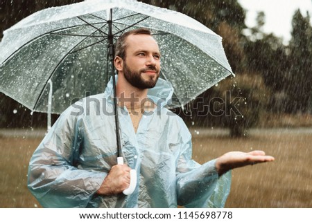 Waist up portrait of smiling male standing outside and holding umbrella. He is stretching hand in delight trying to catch water drops