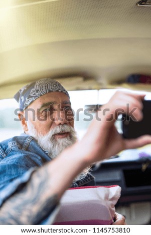 A senior hipster sitting in a car doing a selfie with a phone. he is tattooed and he wears a white beard