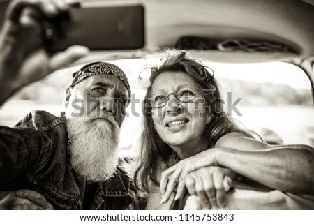 An senior hipster couple sitting in a car doing a selfie with a phone. he is tattooed and he wears a white beard. black and white