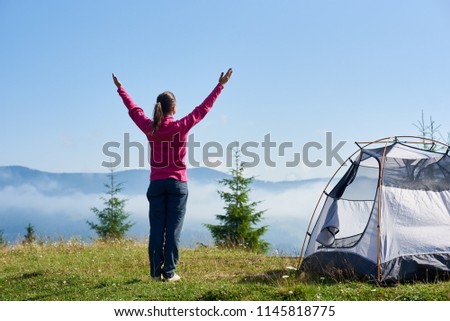 Back view of young long-haired tourist girl standing with raised arms on blooming hill at small tourist tent under beautiful clear blue sky on bright summer morning. Tourism and traveling concept.