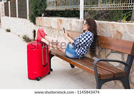 Travel, tourism, technology and people concept - happy woman sit on bench and put her feet on suitcase and typing something on mobile, ready to travel