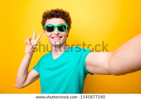 Self portrait of handsome curly-haired glad young guy wearing casual green t-shirt and colorful sun glasses, showing v-sign. Isolated over yellow background