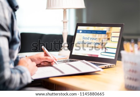 Young man writing college or university application form with pen and paper. Student applying or filling document. Scholarship, grant or admission. School website in laptop.  Royalty-Free Stock Photo #1145880665