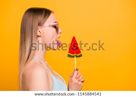 Profile side view of attractive nice cute straight-haired blonde girl, wearing blue swimsuit and color sun glasses, licking candy on stick with tongue. Copy space. Isolated over yellow background