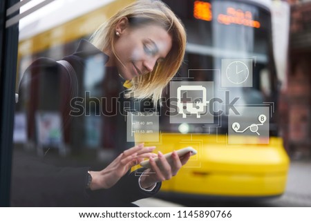Smiling hipster girl waiting for public transport on bus stop while using application on smartphone for checking train schedule. Infographic illustration bus sign. Female chatting on cellular Royalty-Free Stock Photo #1145890766