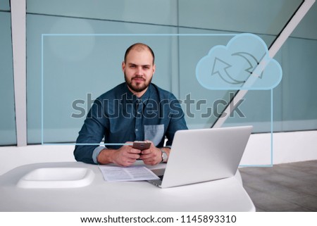 Male IT developer sitting with mobile phone with laptop computer in modern office interior, freelancer guy downloading files to online cloud storage to free up space on netbook. Infographics symbol