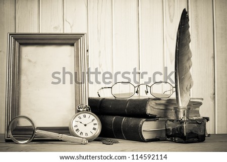 Vintage pocket watch, photo frame, quill and inkwell, books magnifying glass, specs on a table in front of wooden background
