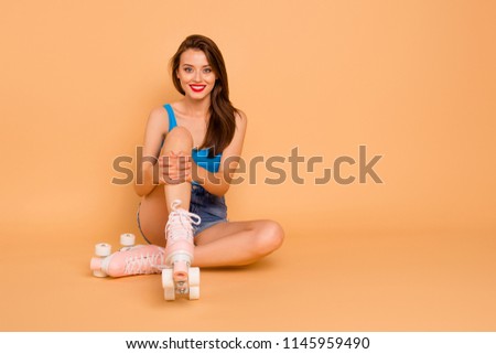 Full-size portrait of creehful and cute sweet girl sits on the floor in roller skates and hugging her knees looking at camera isolated on pastel peach background with copy space for text