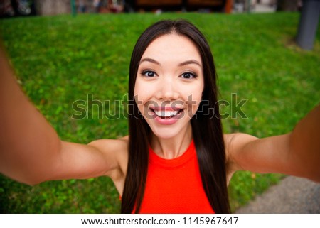 Young korean chinese girl in red dress makes selfie on the camera of a smartphone on the background of a city park