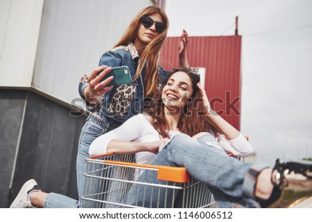Two young happy women having fun shopping trolley race outdoors and make sephi