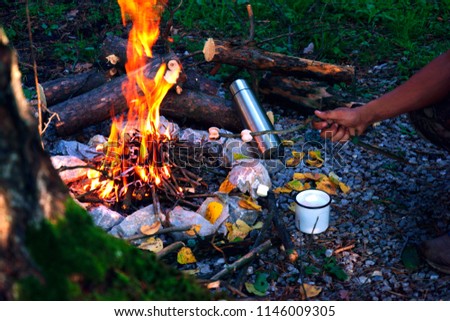 camping outdoors in the fall with a bonfire and coffee in white enamelled iron mug with a thermos and yellow leaves burning fire 