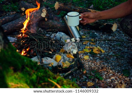 camping outdoors in the fall with a bonfire and coffee in white enamelled iron mug with a thermos and yellow leaves burning fire and marmelo