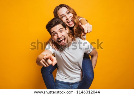 Image of caucasian man giving piggyback ride to joyful woman and pointing fingers at you isolated over yellow background