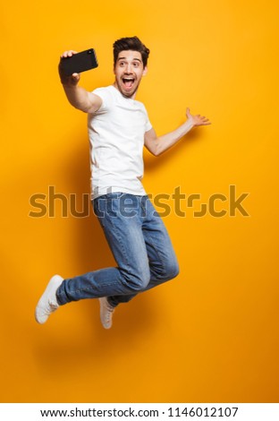 Full length portrait of happy man with brown hair jumping and screaming while taking selfie on black smartphone isolated over yellow background
