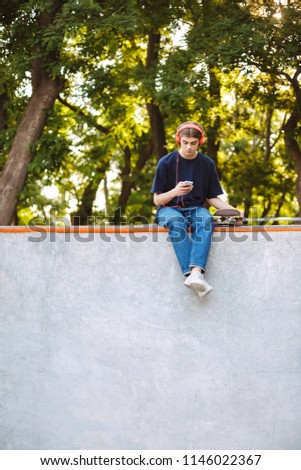 Young cool guy in orange headphones dreamily using cellphone with skateboard near spending time at modern skatepark
