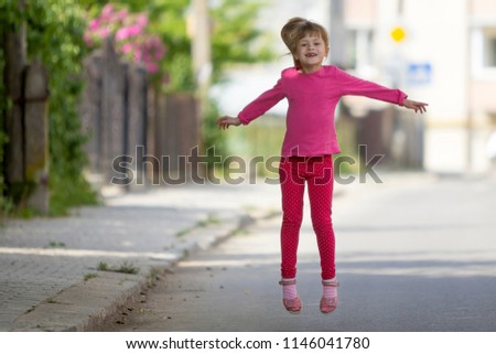 Cute small funny smiling toothless girl in pink casual clothing with long blond pony tail jumping and having fun on blurred bright sunny street outdoors background. Happy careless childhood concept.