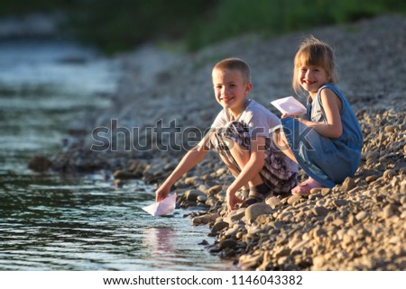 Two cute blond children, boy and girl on river bank sending in water white paper boats on bright summer blurred blue background. Joys and games of happy childhood and outdoors activities concept.