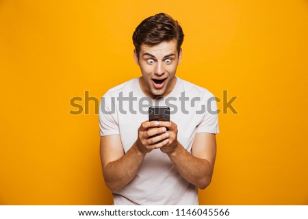 Portrait of a shocked young man looking at mobile phone isolated over yellow background, celebrating