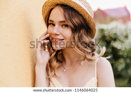 Inspired caucasian woman with shiny wavy hair playfully looking to camera. Outdoor photo of amazing blonde girl in trendy hat.