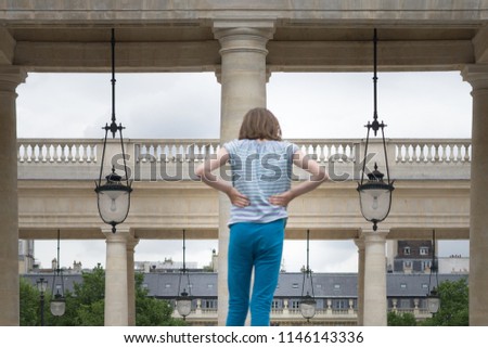 Close up of an anonymous little fair hair girl, hands on hips, back to camera, with fancy parisian landscape, its roofs, columns and old street lamps in background, Palais Royal garden, Paris, France.