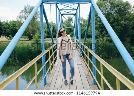 Hipster girl in cool glasses posing for model photo. Young fashionable woman is standing on the bridge. Summer photoshoot in the nature outdoors. Lifestyle concept.