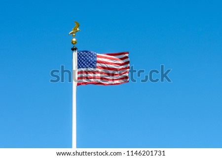 The flag of the United States of America blowing in the wind at full-mast on a white pole topped with a golden eagle on ball against blue sky.